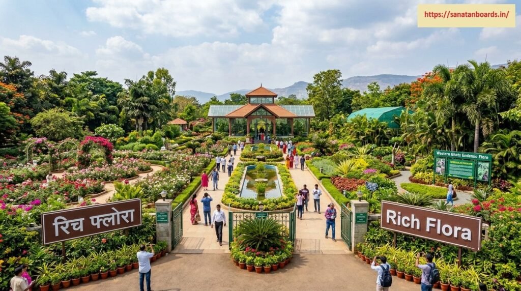 रिच फ्लोरा An elevated view of the entrance and landscaped gardens of Rich Flora, showing visitors strolling along paved pathways lined with vibrant flower beds, water features, potted plants, and a central greenhouse pavilion against a mountain backdrop.
